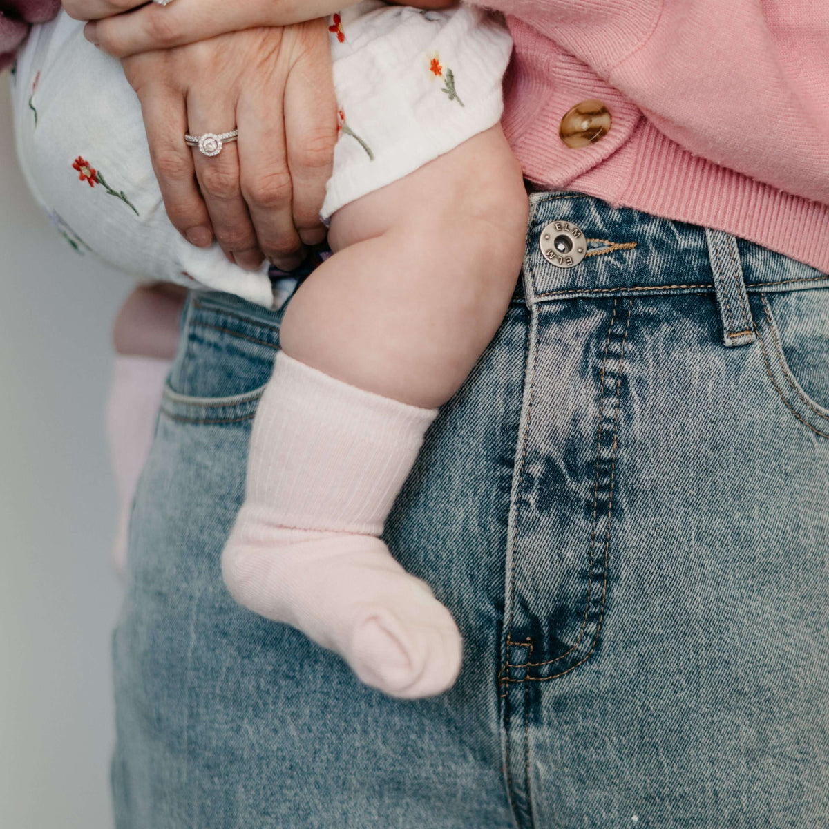 A close-up of a baby wearing blush Organic Cotton Baby Socks while being held by an adult in denim jeans.